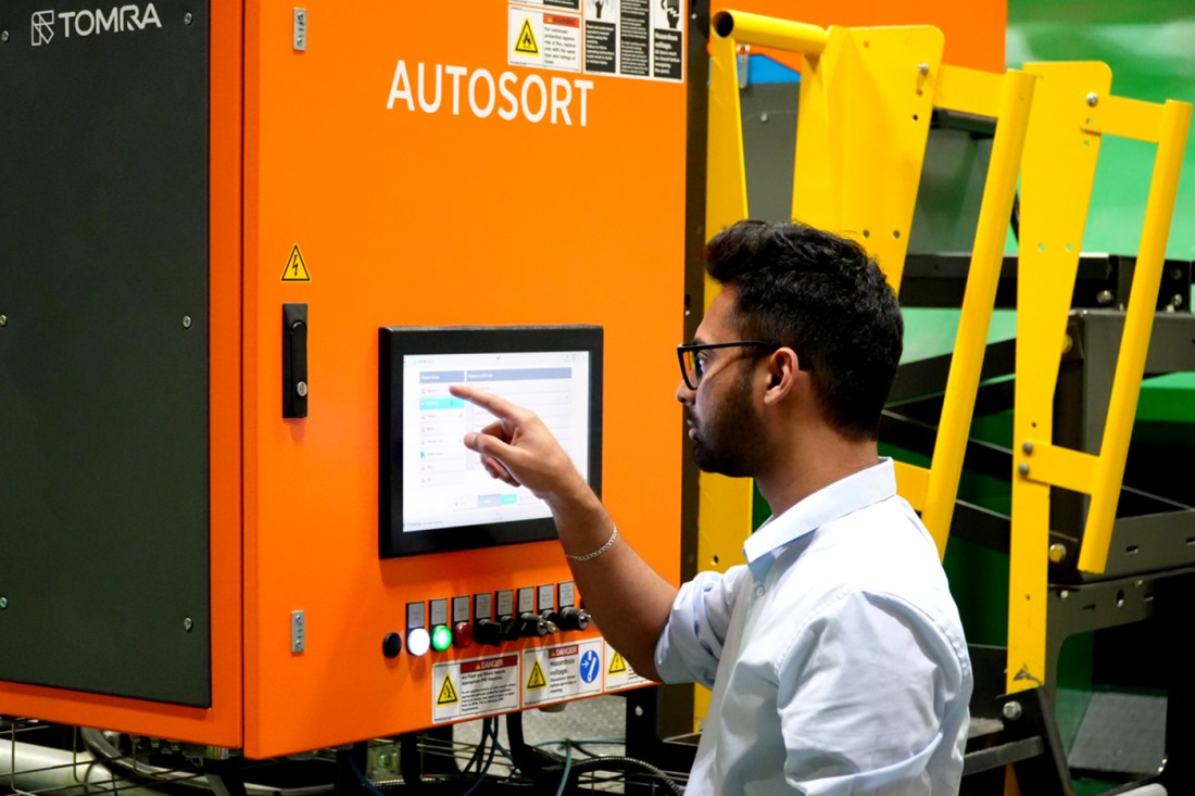 A man with dark hair and glasses points at a touch screen attached to a larger orange machine in a manufacturing plant.