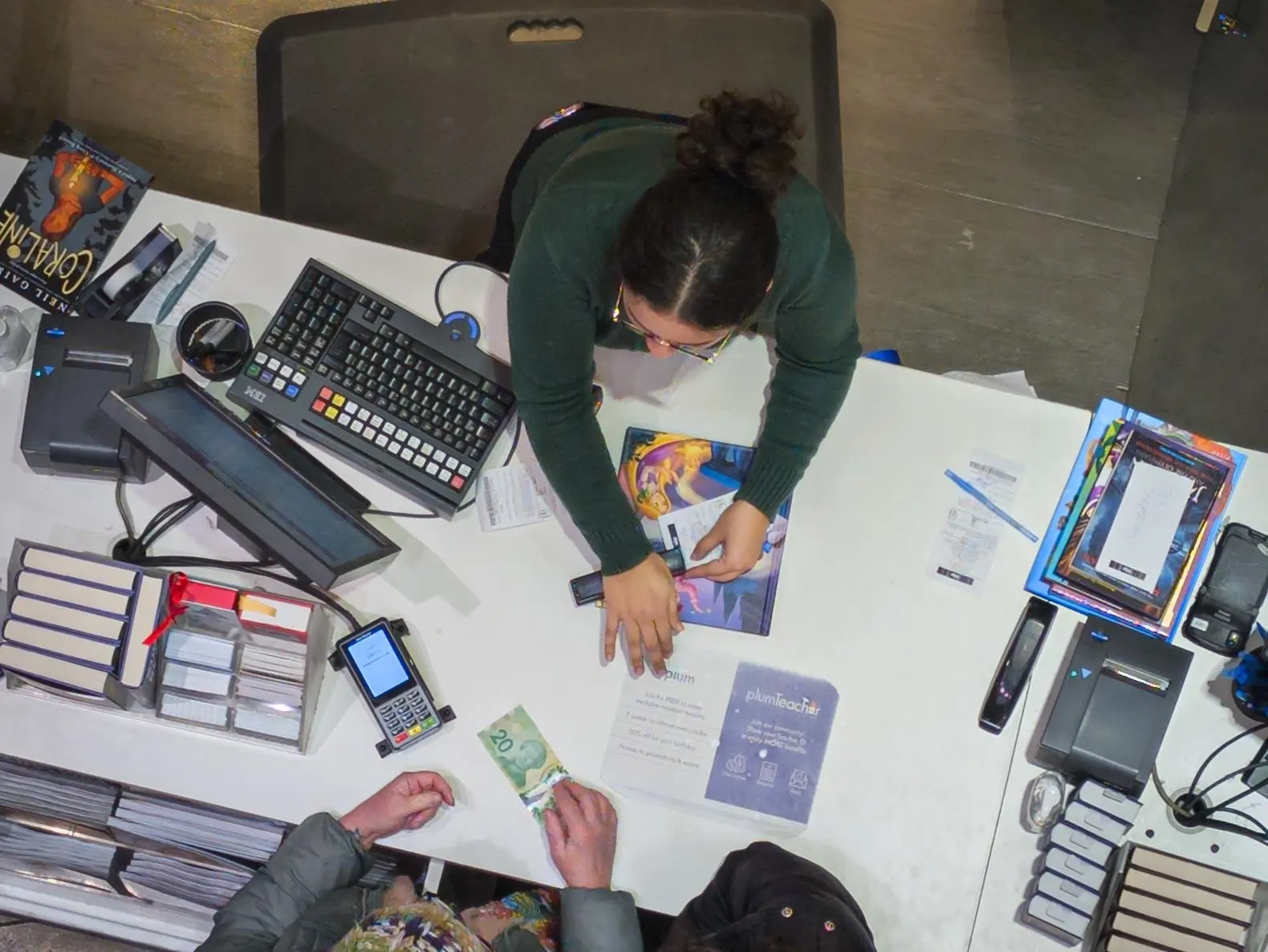 Brightly lit cash register counter with a female cashier and a customer exchanging money at a retail store, diverse items and digital payment devices visible, capturing everyday shopping in Toronto.