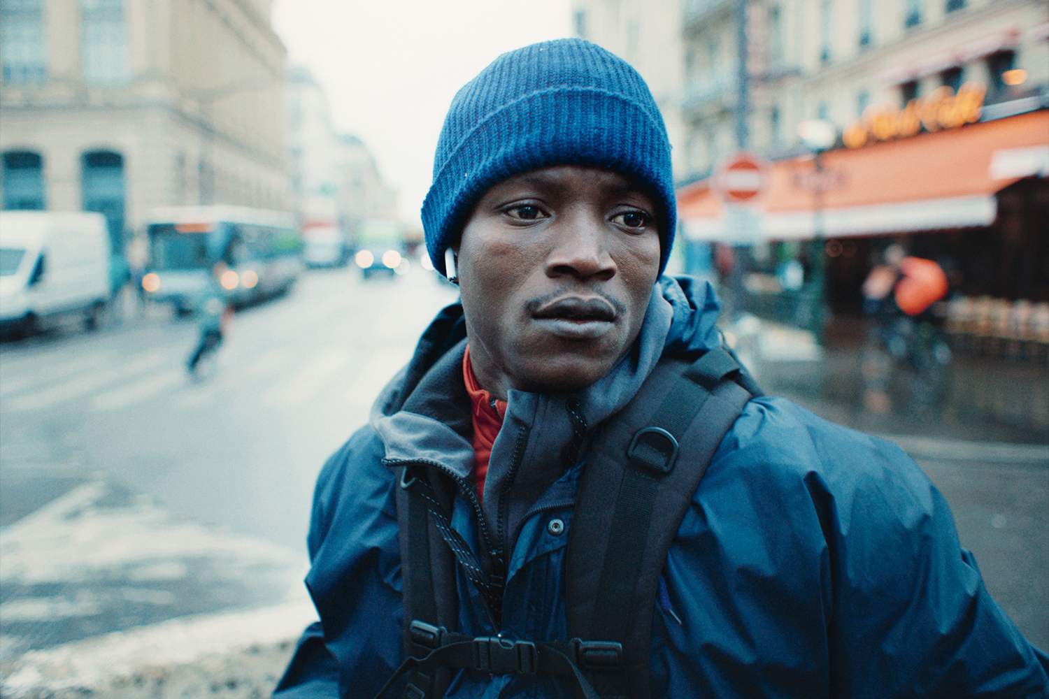 A man wearing activewear, a backpack, and a blue knit hat stands on a corner of a city street and gazes past the camera.