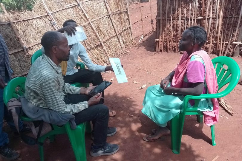 A data collection exercise in Ethiopia, showing a researcher seated outdoors interviewing a refugee woman. They sit on green plastic chairs beside traditional thatched fencing, with the researcher using a tablet and holding a form during the discussion.