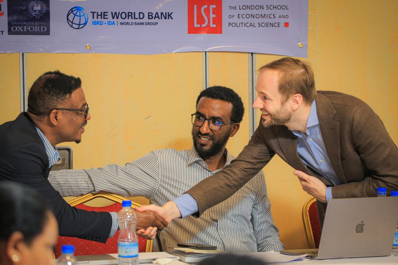 Future of Development director at a workshop with Ethiopian partners, shown shaking hands with a participant while another looks on. A banner with partner logos hangs behind them, and a laptop and water bottles sit on the table.