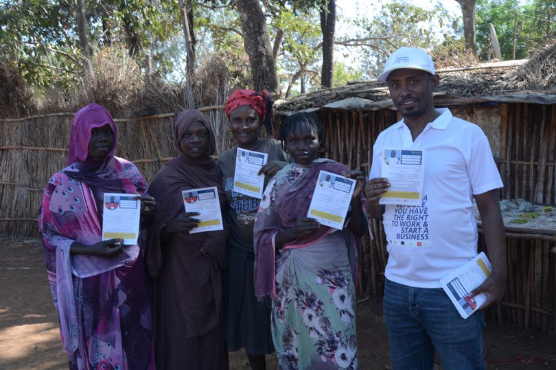 A group of five adults standing outdoors in front of a rustic wooden structure in Ethiopia, holding up work permit documents issued through a refugee support programme. The individuals wear a variety of brightly coloured clothing and headscarves, and one man on the right wears a white cap and polo shirt. They face the camera and display the permits proudly, suggesting successful participation in the programme.