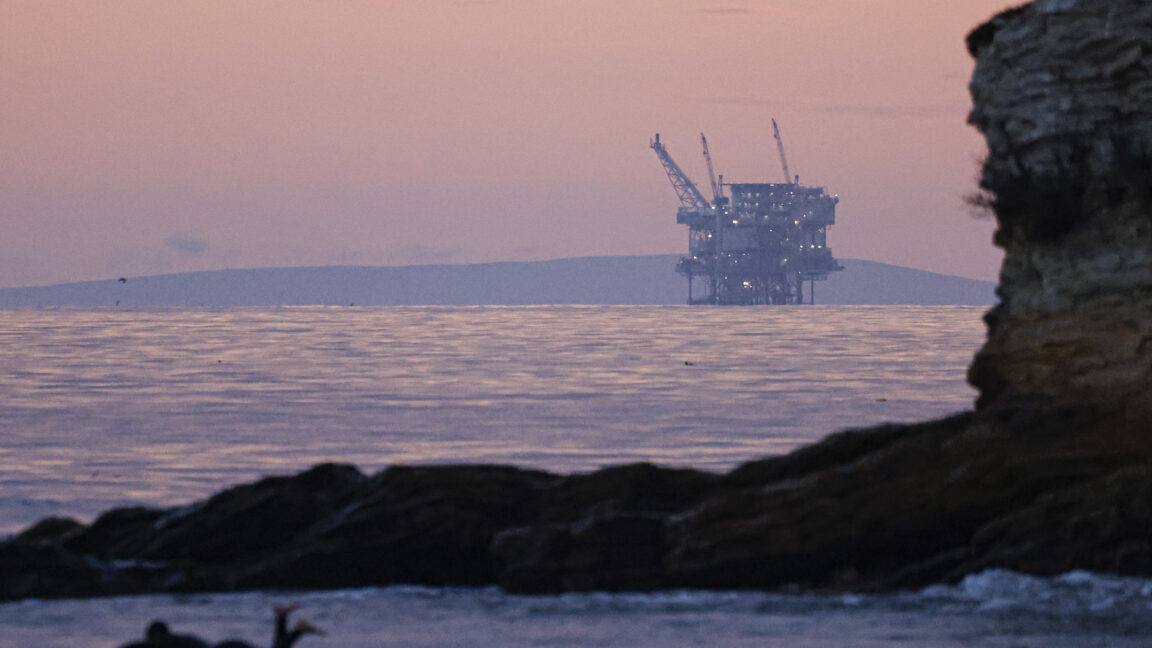 A surfer is paddling in the ocean water as the Hondo offshore oil platform looms in the distance at Refugio State Beach Santa Barbara, California.