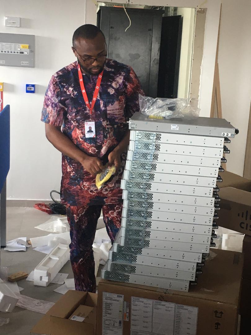 A man in a patterned shirt, pants, and glasses stands next to a stack of Cisco networking equipment, holding cables, in an IT installation environment.