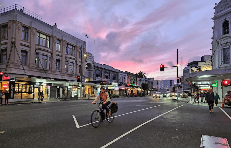 A person rides a bicycle through a city intersection at dusk, passing shops and restaurants with glowing signs under a pink and purple sky. Cars and pedestrians are visible in the background, and traffic lights are red.