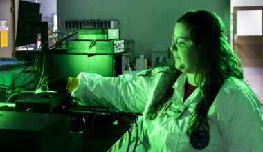 A scientist wearing safety glasses, gloves, and a protective lab coat operates equipment in a laboratory illuminated by green light. She reaches toward an instrument surrounded by cables and electronic devices, concentrating on her work in the controlled lab environment.