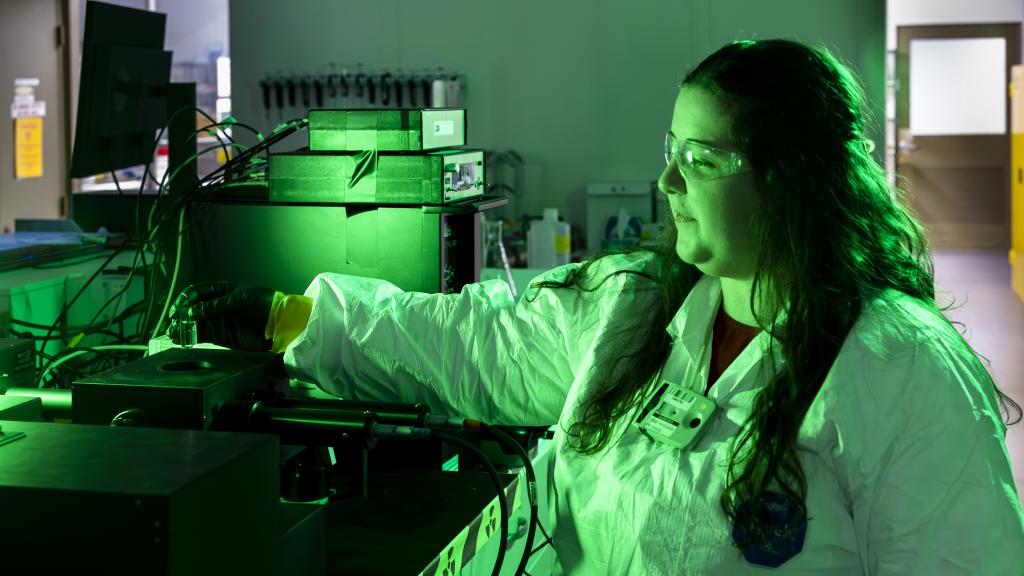 A scientist wearing safety glasses, gloves, and a protective lab coat operates equipment in a laboratory illuminated by green light. She reaches toward an instrument surrounded by cables and electronic devices, concentrating on her work in the controlled lab environment.