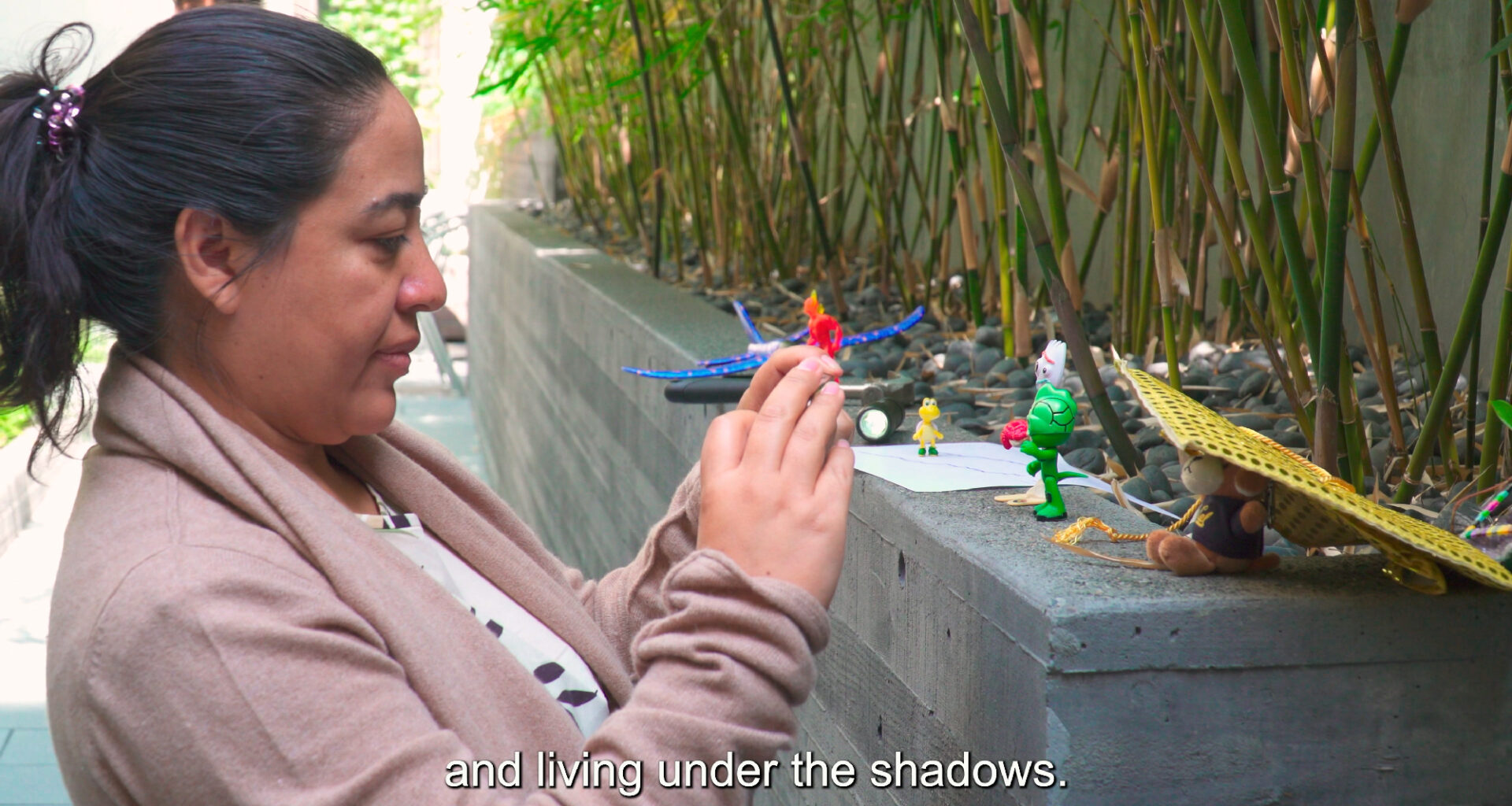 person holds phone up while creating video using small toys, subtitle reads 'and living under the shadows.'