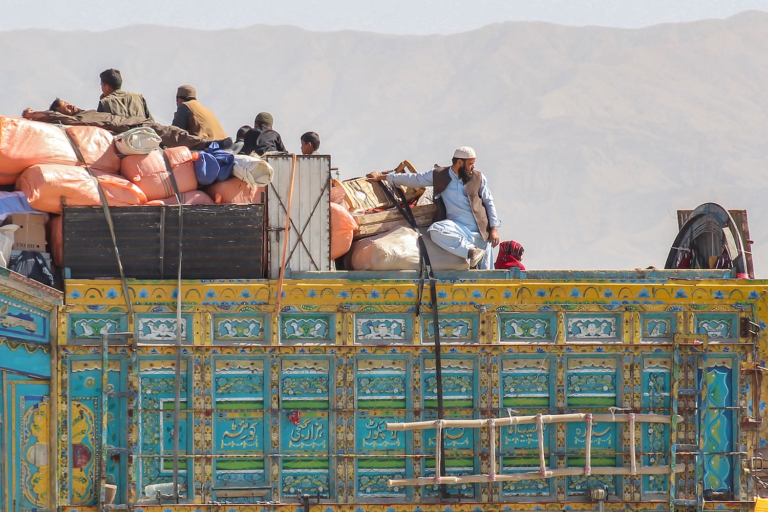 About half a dozen people perch on the back of a truck piled high with bundles and trunks. One man at the back has his hand splayed over a suitcase as he looks over his shoulder. A mountain looms against a hazy sky in the background.