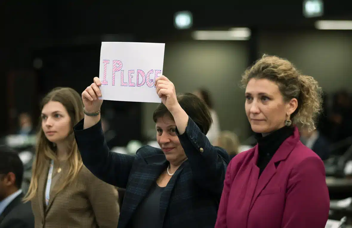 Three women are seen in the photo with the one in the middle holding up a piece of 8 by 11 paper with "I pledge" written on it by hand.