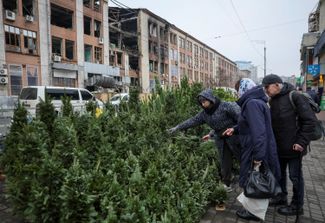 People shop for Christmas trees at a street market in front of a building damaged by Russian missile and drone strikes. Kyiv, December 16, 2025.