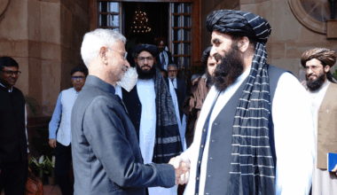 India External Affairs Minister Dr. S. Jaishankar and Minister of Foreign Affairs of Afghanistan, Amir Khan Muttaqi shake hands outside a building. Jaishankar wears a grey suit, Muttaqi a black turban and waistcoat over a white shirt with striped scarf.