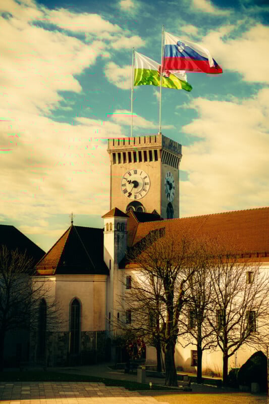 A clock tower with three flags, including the Slovenian flag, rises above a historic building with red-tiled roofs and leafless trees on a partly cloudy day.