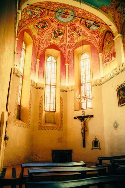 Small chapel interior with wooden pews, tall arched windows letting in sunlight, a crucifix on the wall, ornate painted ceiling with religious artwork, and a chandelier hanging near the altar.