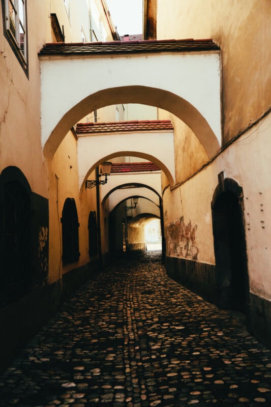 Narrow cobblestone alleyway with arched bridges connecting old, beige buildings; black metal lantern hangs from the wall. Sunlight glows at the end of the passage, suggesting an opening ahead.