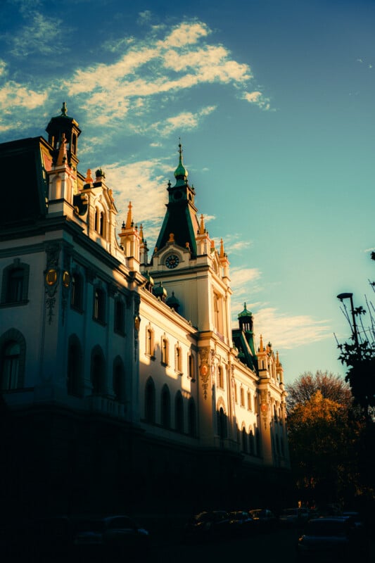 A grand historic building with ornate towers and a clock, bathed in warm sunlight against a blue sky with scattered clouds. Trees and parked cars line the street below.