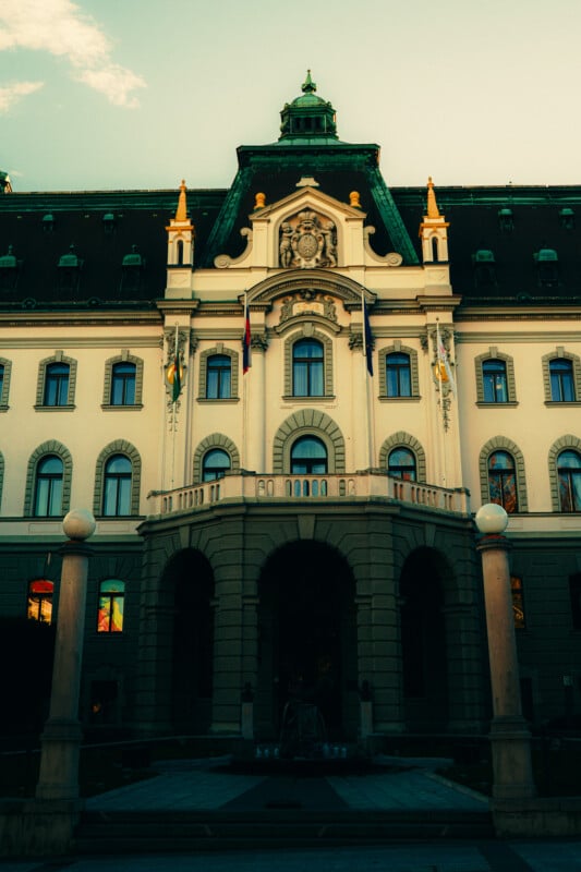 A grand historic building with arched entrances, ornate decorations, and a green-tiled roof, photographed at dusk with a moody sky in the background. Pillars frame the entrance in the foreground.