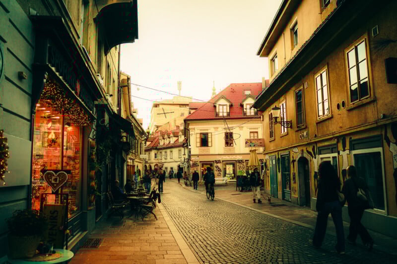 A charming European street at sunset, lined with colorful buildings, shopfronts, and outdoor seating. People stroll and cycle along the cobblestone path, creating a lively and inviting atmosphere.