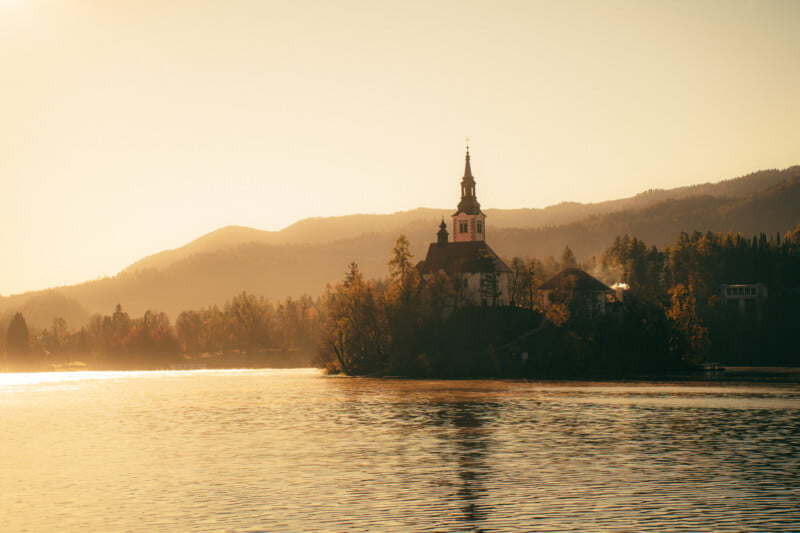 A church with a tall steeple stands on a small wooded island in a lake, surrounded by calm water and misty mountains in soft golden sunlight.
