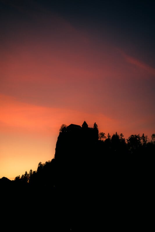 A dark silhouette of a castle on a hilltop is set against a vibrant sunset sky, with shades of orange, pink, and purple blending above the horizon. Trees surround the hill, enhancing the dramatic scene.