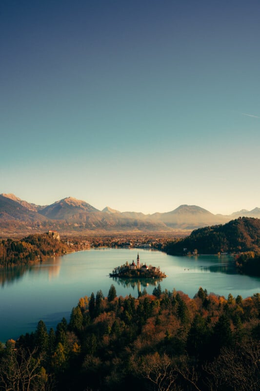 A scenic view of Lake Bled in Slovenia, featuring a small island with a church in the center, surrounded by calm blue water, lush forests, and distant mountains under a clear sky.