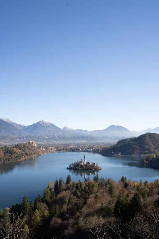 A small island with a church sits in the middle of a blue lake, surrounded by forested hills and distant mountains under a clear blue sky.