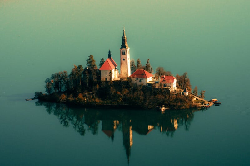 A small island with a church and several buildings sits in the middle of calm, blue-green water, reflecting the structures and surrounding trees.