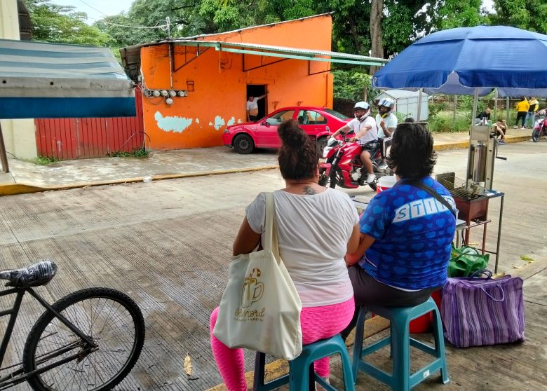 Two people on stools on the sidewalk of a road where a motorbike with two people on it is crossing, sitting across the street from colorful buildings