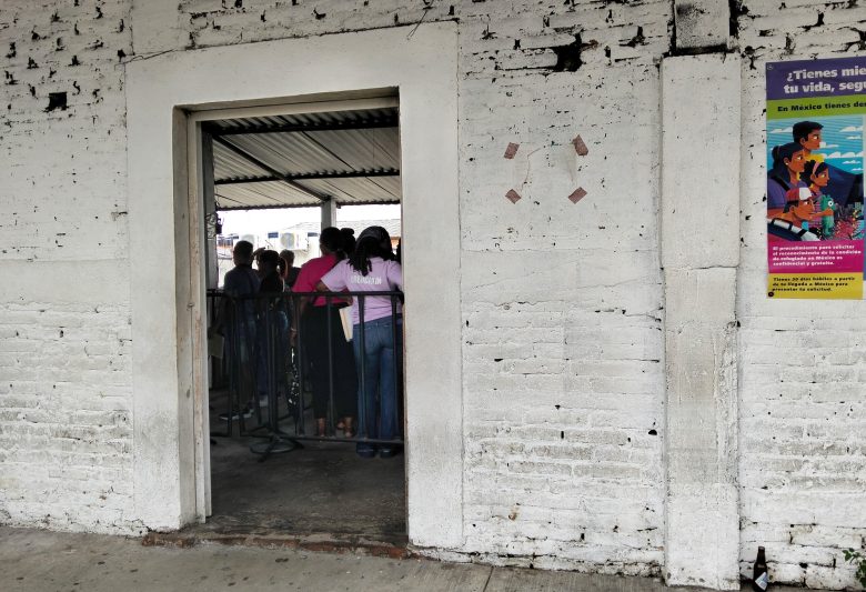 People standing inside a sheltered area seen through a doorway in a white painted brick wall with a poster on the side.