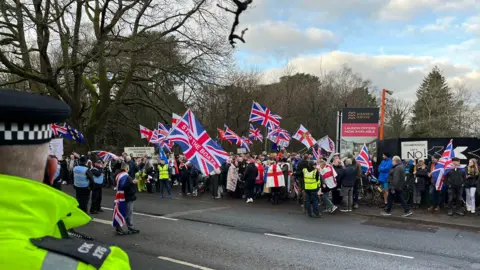 A police officer in hi-vis jacket watching a group of protesters marching along a road. They are holding Union Jack and St George flags.
