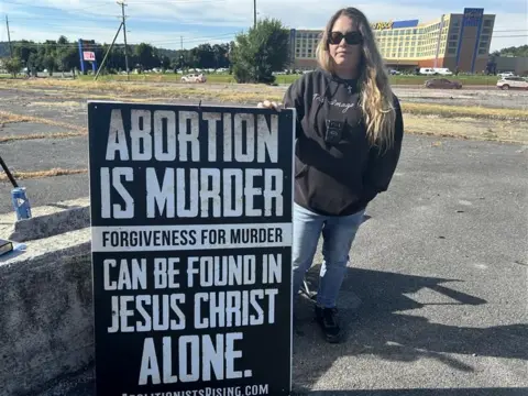 A woman stands in a parking lot with a sign that says: Abortion is murder, forgiveness for murder can be found in Jesus Christ alone.