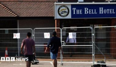 Two men in T-shirts and shorts walking with their backs to the camera towards The Bell Hotel.  There is a metal fence in front of the hotel building.