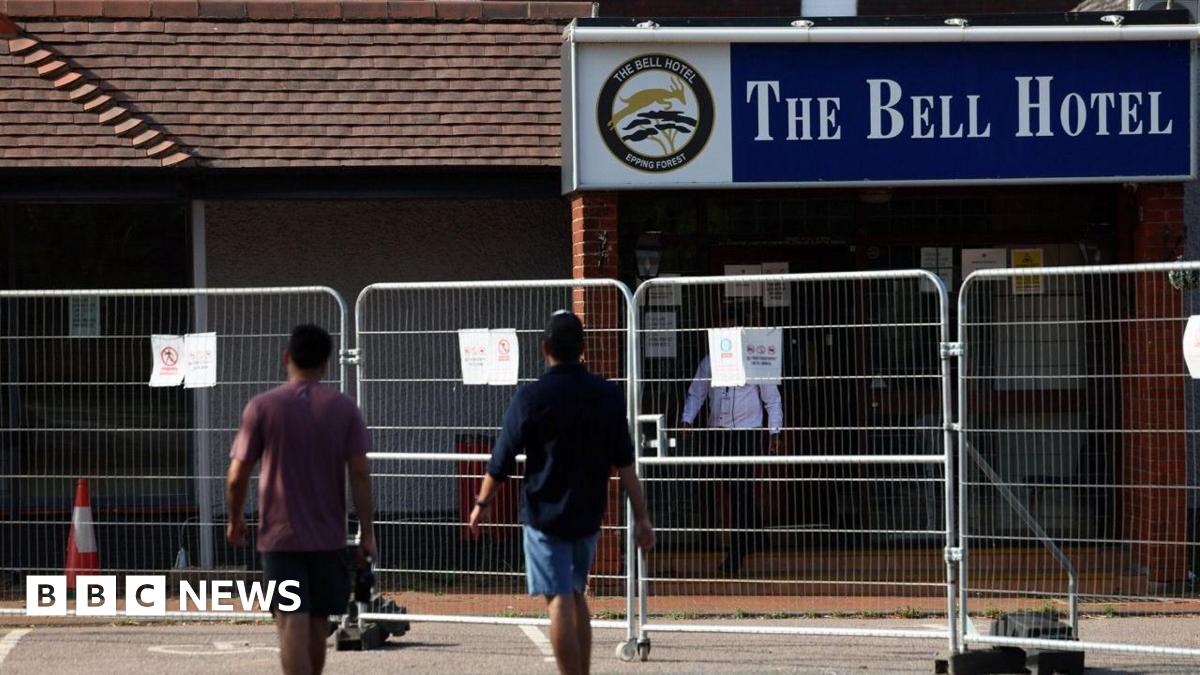 Two men in T-shirts and shorts walking with their backs to the camera towards The Bell Hotel.  There is a metal fence in front of the hotel building.