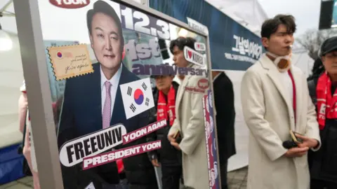BBC/Hosu Lee A mirror with a large sticker of former South Korean President Yoon Suk Yeol, alongside other stickers, including ones that say "FREEUNIV", "YOON AGAIN", "PRESIDENT YOON", and "1st anniversary". Two people stand alongside it, one of them wearing a long white coat