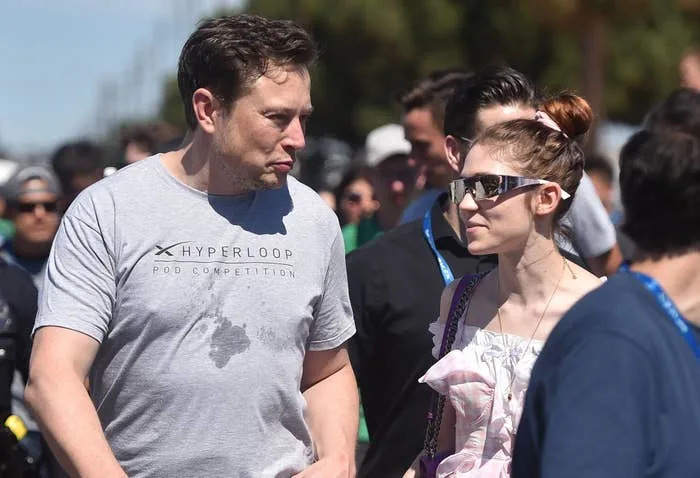 Elon Musk wears a casual logo t-shirt; Grimes wears reflective sunglasses and a ruffled top, walking outdoors