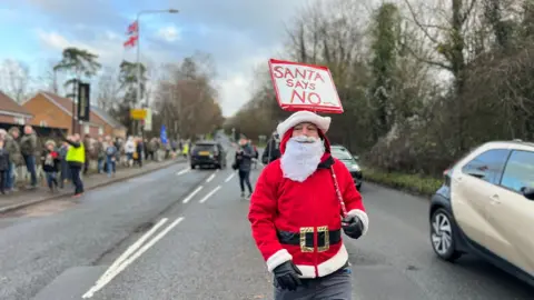 A man dressed in a Father Christmas holding a sign which reads "Santa says no". There is a beige and black car driving past him and some protesters on the other side of the road.