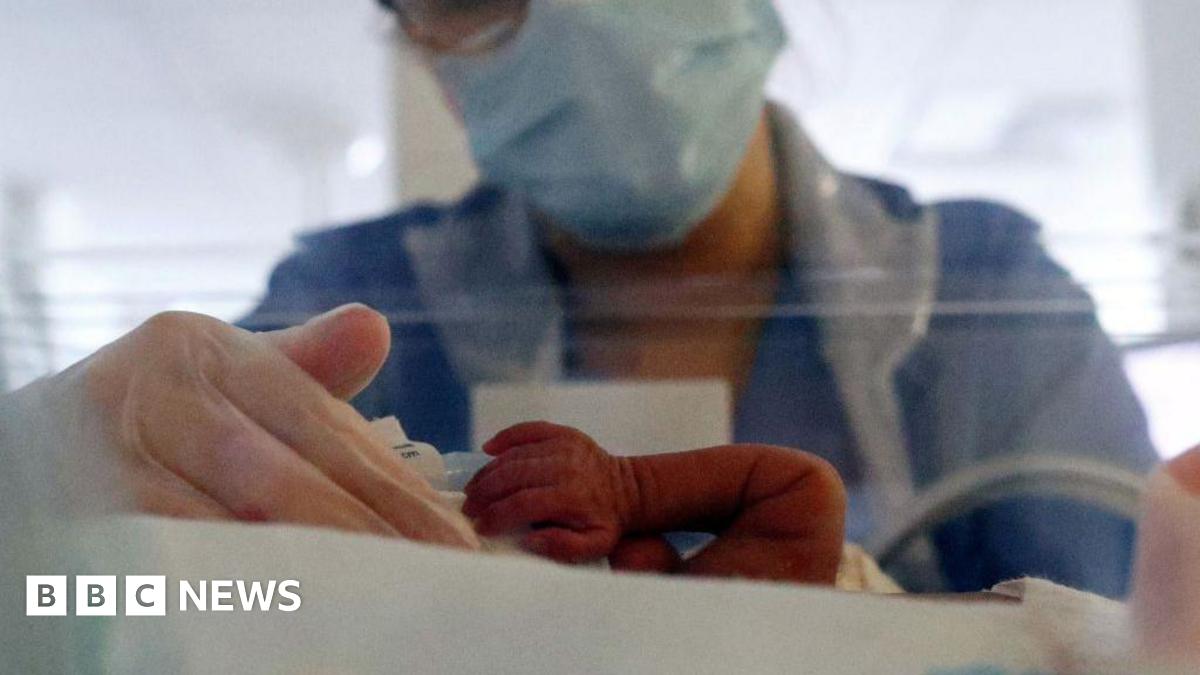 Baby in neonatal ICU incubator, with only an arm visible. A nurse with a face mask, gloves and glasses, has her hand curled around the baby