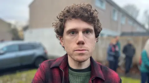 Neil Hepburn has brown curly hair. He is wearing a red checked jacket over a green jumper.