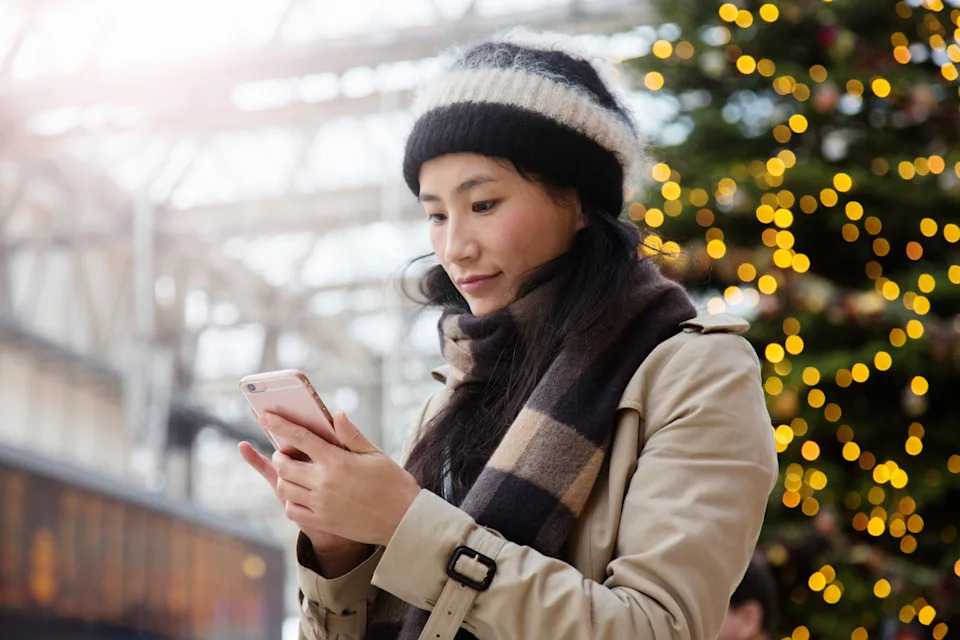 Woman looking at her phone while standing in front of Christmas tree. (Getty Images)