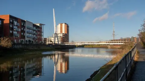 Getty Images The cable-stayed 'Sir Bobby Robson' pedestrian footbridge over the River Gipping, Ipswich on a sunny day. Large blocks of flats line the left hand side of the river while there is a footpath running on the right hand side. 