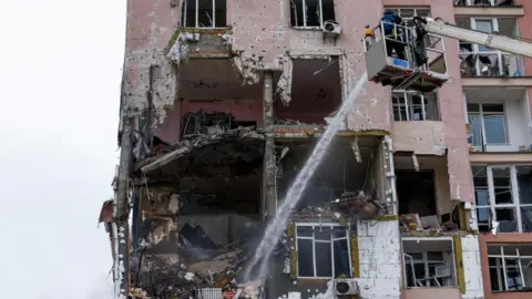Reuters A pink apartment building with a huge hole in the side of it and a firefighter on a crane spraying water into it.
