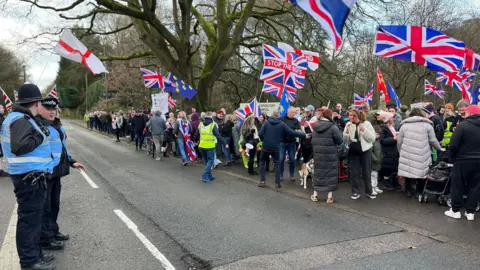 A line of people standing on a road and holding Union Jack and St George flags.