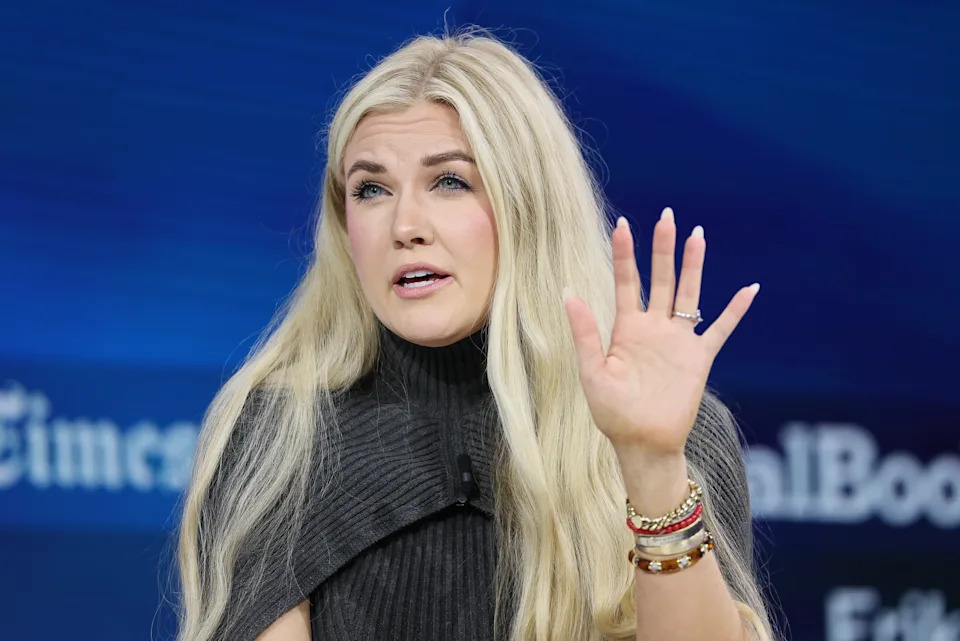Person with long hair gestures while speaking at a news event, wearing a textured high-neck top and several bracelets