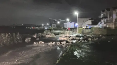 A number of large rocks near the seawall are visible scattered along a footpath. 
A number o street lights and buildings can be seen in the background