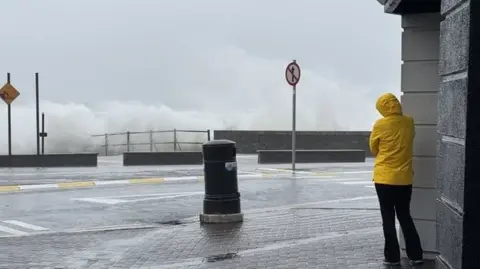 RTE A woman, wearing back trousers and a yellow anorack pulled tight, stands with her hood up, faces a thick spray that is coming off heavy waves crashing into the quay. A black street bin and about twenty yards of footpath and tarmac separate the woman and the tumultuous ocean. 