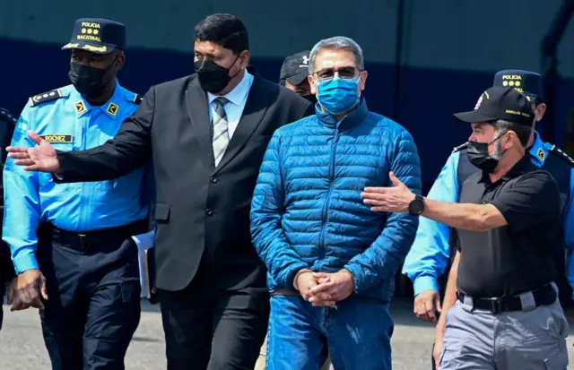 Juan Orlando Hernández vistiendo una chaqueta deportiva azul, pantalones vaqueros y una mascarilla azul camina con las dos manos juntas puestas al frente, rodeado de varios efectivos policiales.