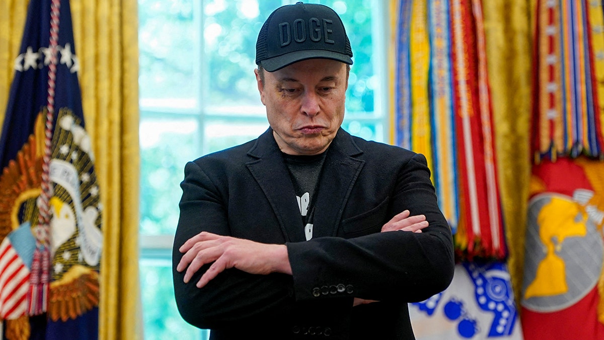 Elon Musk looks down, standing in the Oval Office with his arms crossed