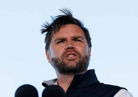 jd vance speaking at a podium with a U.S. emblem, wearing a casual zip-up vest and shirt outdoors