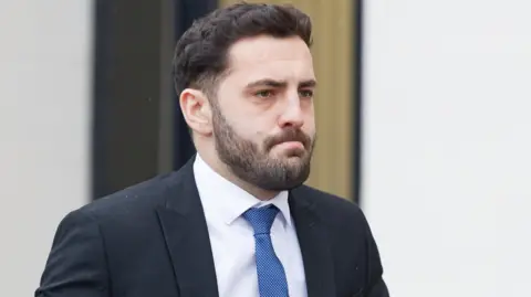 Athena Pictures Agency A man is wearing a black suit jacket, a blue tie and a white shirt. He has a beard and black hair.