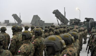 Żołnierze w pełnym umundurowaniu stoją w szeregu przed wyrzutniami rakiet Patriot.